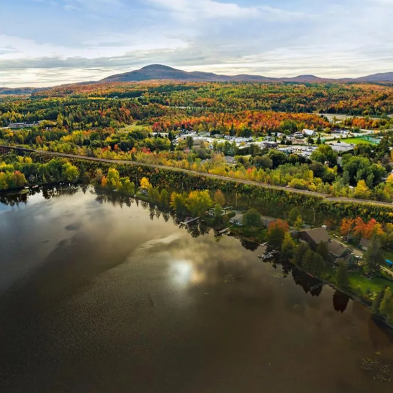 Vue aérienne d'Eastman à l'automne : lac bordé d'érables rouges et orangés, maisons du village le long de la rive et montagne à l'horizon