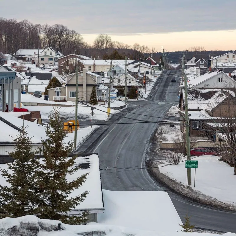 Rue de village enneigée à Saint-Benoît-Labre : maisons et toits couverts de neige de part et d'autre de la chaussée, épinettes au premier plan