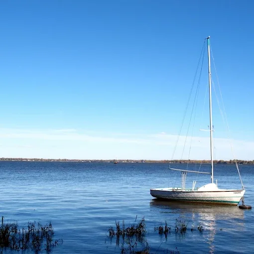 Voilier au mouillage sur un grand lac calme, sous un ciel bleu dégagé, quenouilles au premier plan