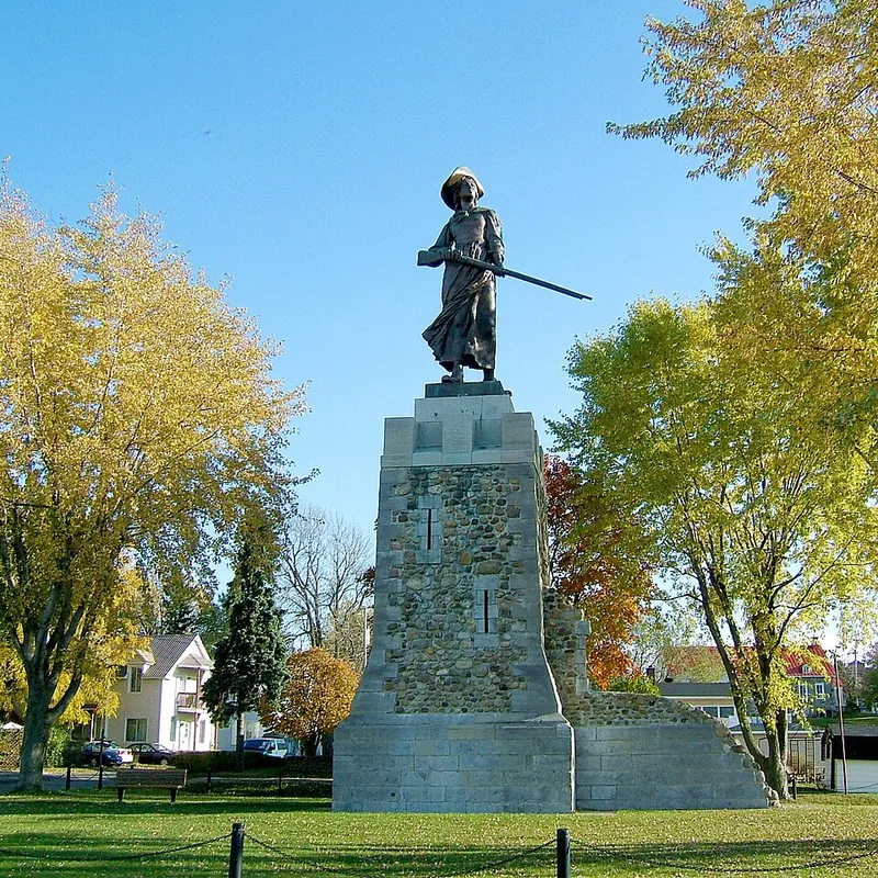 Monument de Madeleine de Verchères : statue de bronze sur un haut socle de pierre, dans un parc aux arbres jaunis par l'automne